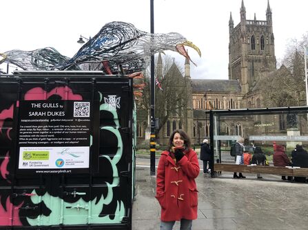 A photo of a lady with brown hair wearing a red duffel coat standing next to a plinth with a seagull sculpture on top in the middle of a city centre with a cathedral behind