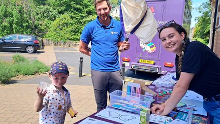 Community outreach stall: a smiling woman leans over a table of coloring pages, markers and jars while a man holds an insect net and a young child shows a clear container and sticker beside a purple outreach van.