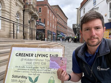 Staff member holding a flyer next to a board titled "Greener Living Fair," promoting a sustainable living event with shoppers and red-brick town centre buildings visible in the background.