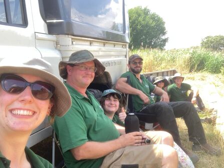 Trainees in green shirts and sun hats taking a shaded break beside a truck in grassy countryside, smiling after outdoor conservation work.