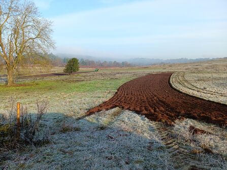 Frost-covered rural field with a curving strip of freshly ploughed dark soil in the foreground, a small tractor working near a lone tree in the midground, a large bare tree at left and a misty treeline beneath a pale blue sky.