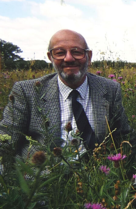 A man in glasses and with facial hair is sitting in a meadow and is smiling at the camera