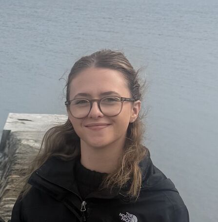 A head and shoulders shot of a woman wearing a black coat and glasses with the sea in the background