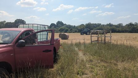 Pickup truck parked at the edge of a harvested field with its door open and a person inside, while round hay bales and a tractor work across the field near a tree line under a bright blue sky.
