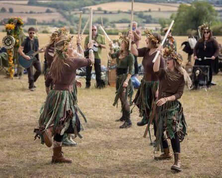 A photo showing a group of Morris dancers in green and brown clothing with leaf crowns dancing in a field holding poles with countryside behind