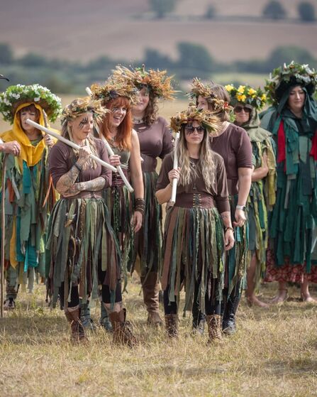 A photo showing a group of Morris dancers in green and brown clothing with leaf crowns standing in a group in a field with countryside behind