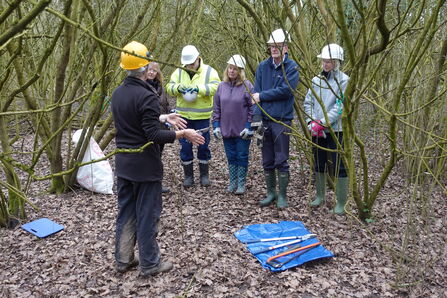 A photo showing six people standing in a winter woodland wearing hard hats and welly boots, with one person standing in front of the group with their hands out explaining something