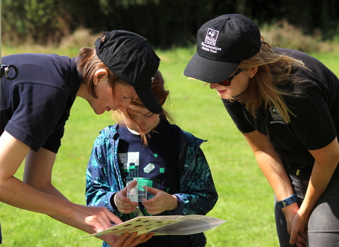 2 young women with WWT logos helping a young girl to identify an insect by Lauren Roberts