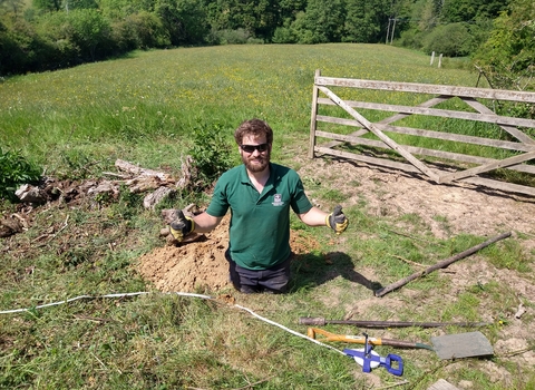 Young man in the middle of a job in a field on a sunny day who's paused and is giving 'thumbs up' to the camera