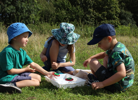 Three children with minibeast pots in a grassy field by Lauren Roberts