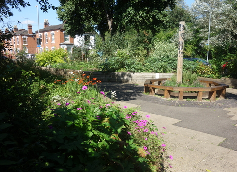A circular bench in the middle of a flower-rich greenspace with houses in the background by Liz Yorke