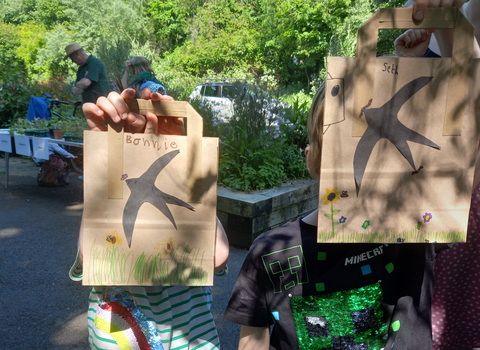 Two children holding up paper bags that they've decorated with swifts and wildflowers by Liz Yorke