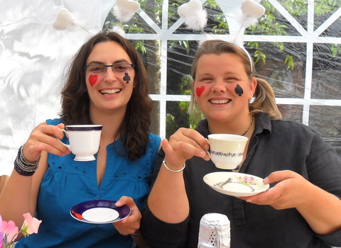 Two women looking at camera and holding up cups and saucers; the women have hearts, clubs and spades painted onto their faces