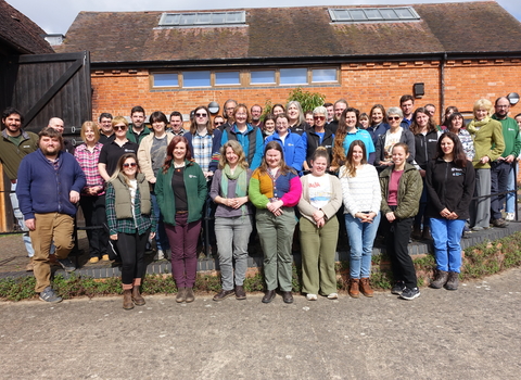 A group of about 40 people smiling at the camera, standing in front of a red brick building