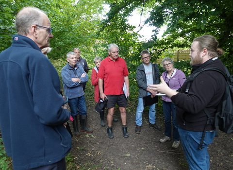 A group of people in a woodland - they're standing in a group looking at and listening to a person with long hair and a beard 