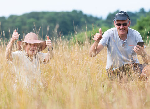 Two volunteers are crouching/sitting in amongst vegetation on a summer day and both are looking at the camera with their thumbs up