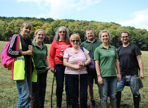 A group of people smiling at the camera - they're in a meadow on a sunny day and are holding tools