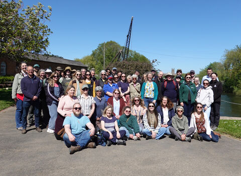A large group of people posing and smiling for the camera - it's a bright sunny day, there's a canal on the right hand side and Worcester Cathedral is just visible in the background